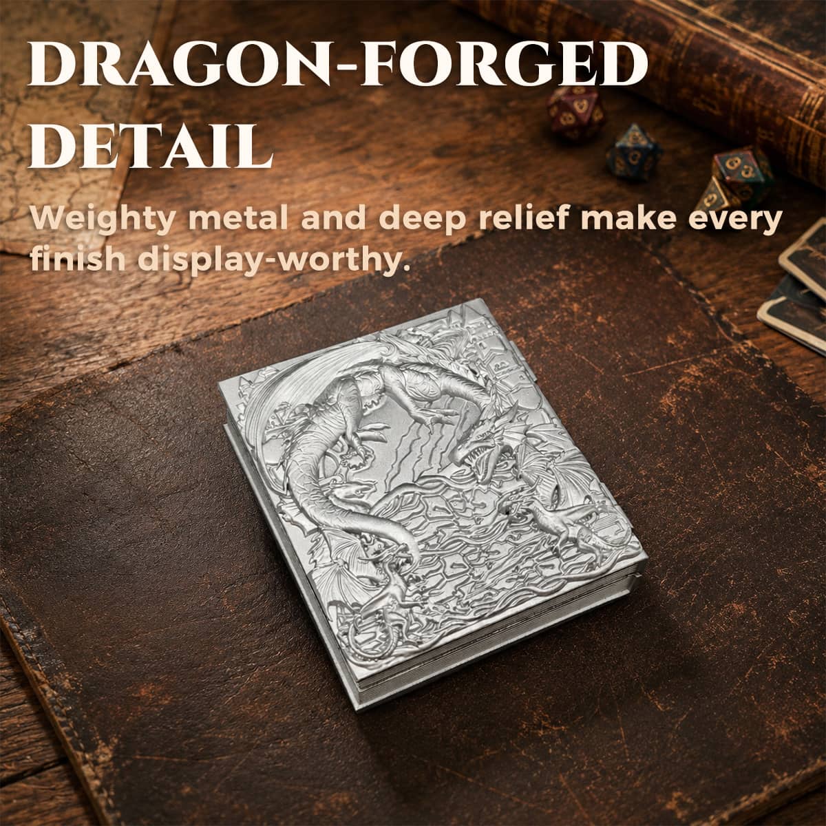 Closed silver metal dice codex with deep dragon relief on a leather tabletop.